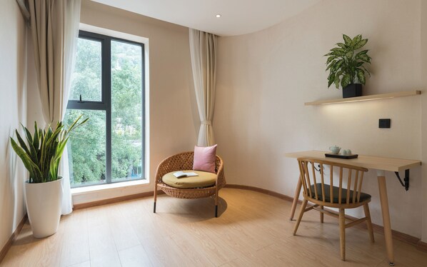Une pièce avec une grande fenêtre, un fauteuil en osier avec un coussin, une petite table avec une plante, et une plante en pot placée près de la fenêtre.