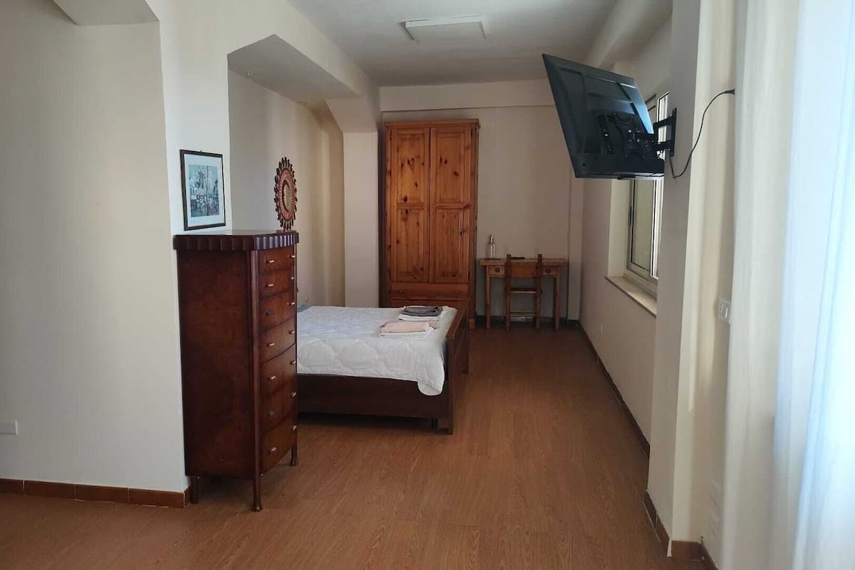 Ein schmaler Flur mit einem Bett, einem Schminktisch, einem Holzschrank, einem kleinen Tisch mit Stuhl und einem an der Wand befestigten Fernseher.