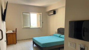Ein Schlafzimmer mit Bett, einem Fenster mit Jalousien, einem an der Wand montierten Fernseher und einem kleinen Holztisch mit Handtuch.