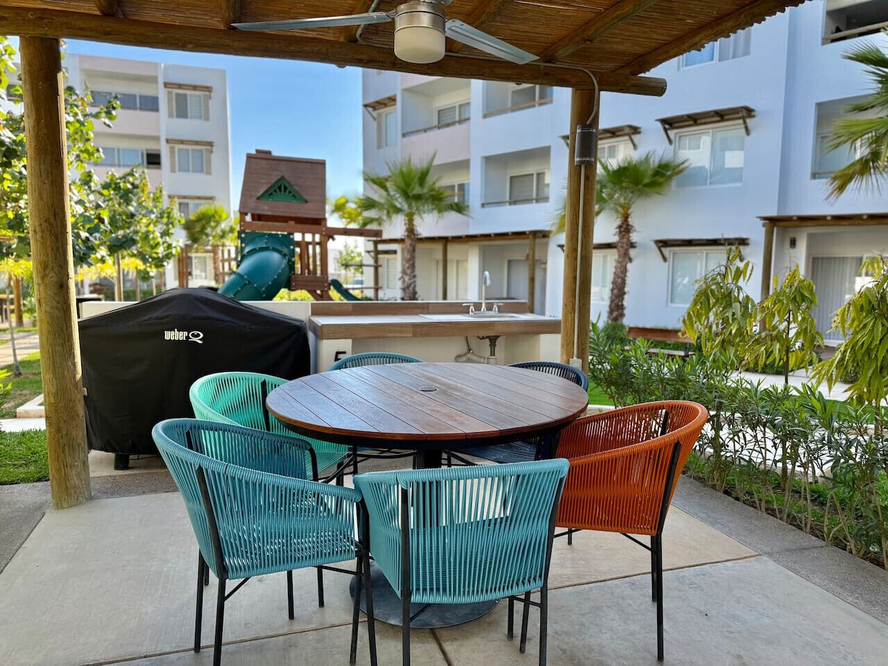 An outdoor patio with a round wooden table and wicker chairs, a barbecue grill, and a view of residential buildings in the background.