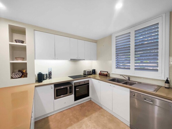 A modern kitchen with white cabinets, a built-in oven, microwave, and dishwasher. There is a window with blinds and a shelf with decorative items. - Villa Amore - Two-storey villa at Cypress Lakes Resort (Pokolbin)