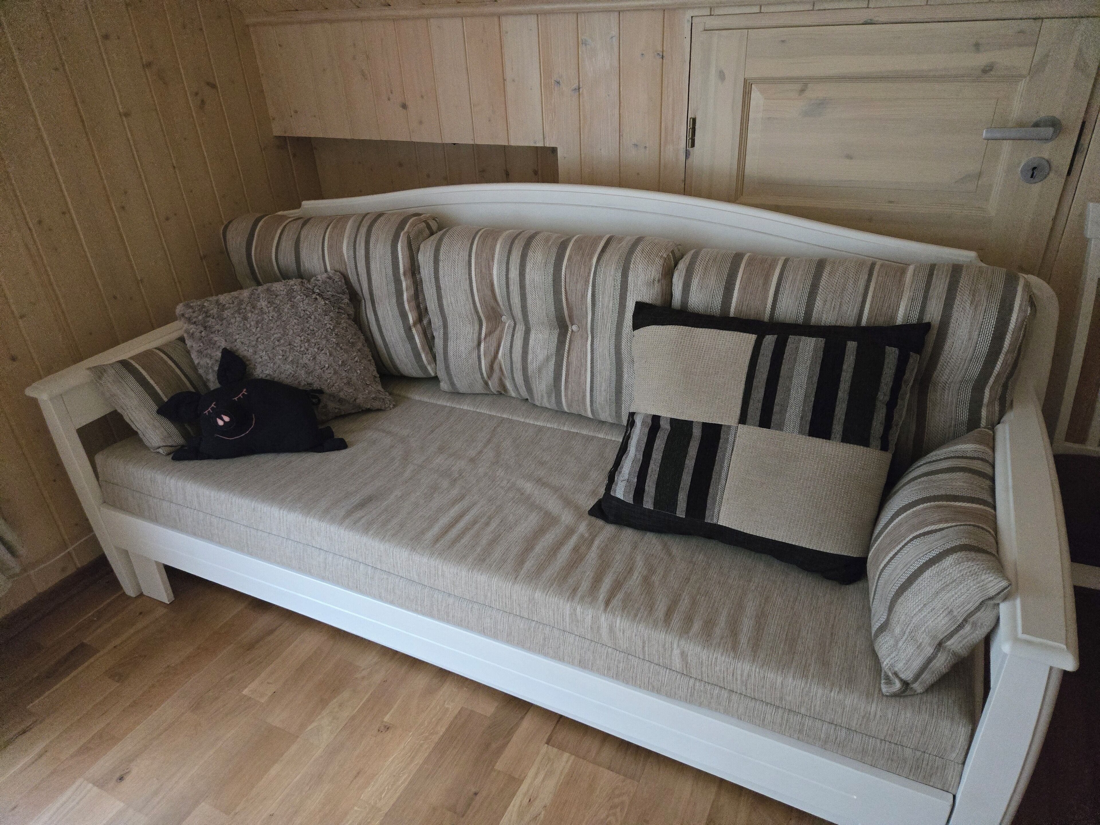 Un sofá blanco con cojines a rayas y un juguete de peluche negro en una habitación con paneles de madera y suelo de madera.