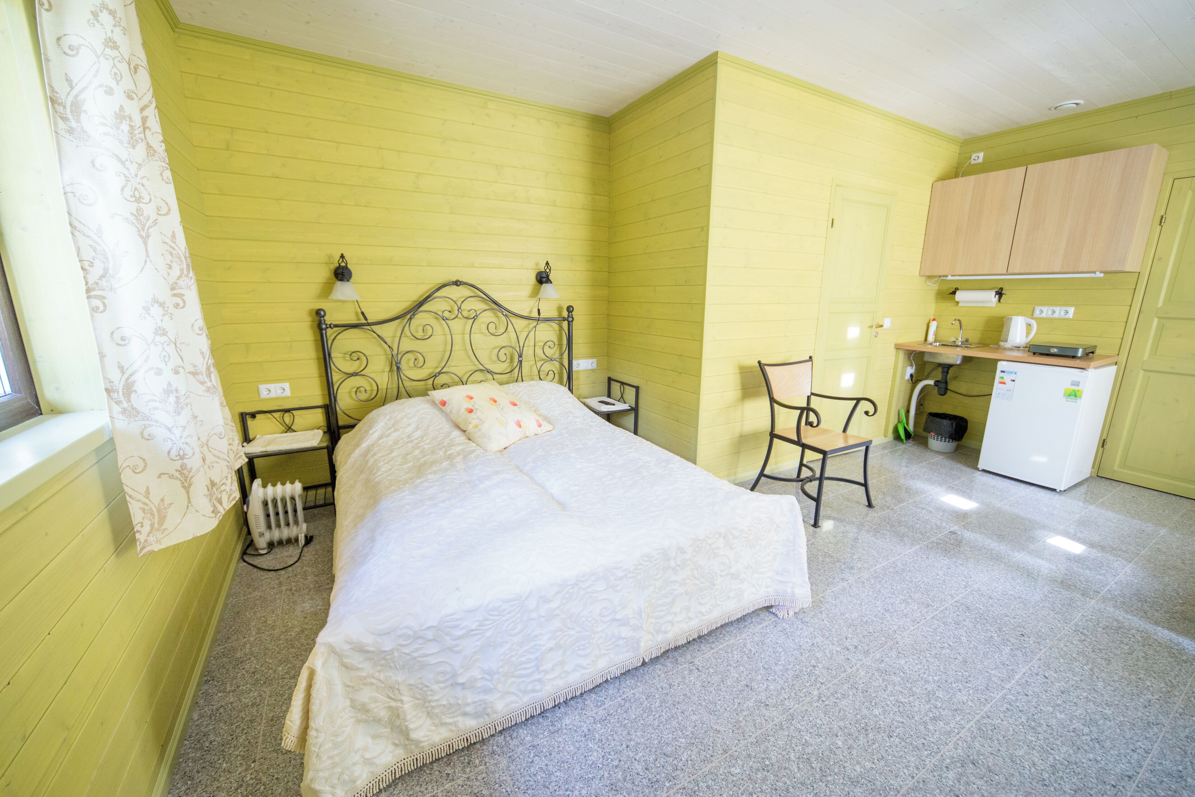 A room with a bed, a small desk, a chair, a radiator, and a kitchenette.