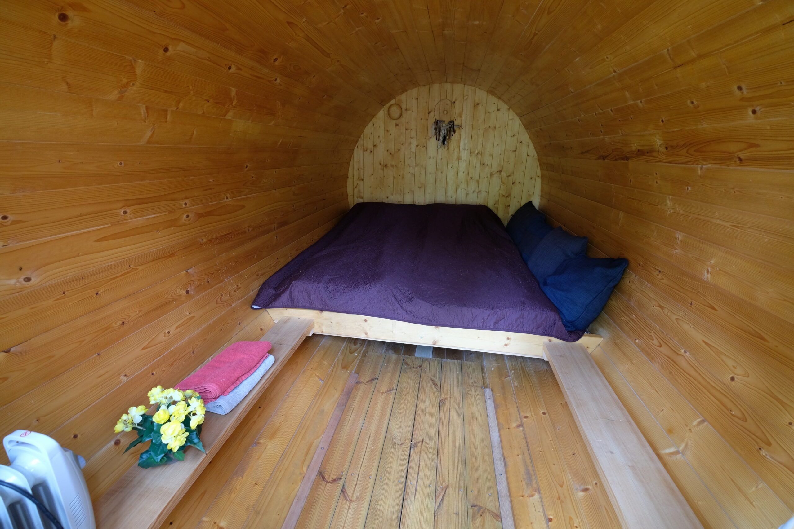 Un interior de madera con una cama, almohadas, una mesita con una toalla rosa y un ramo de flores.