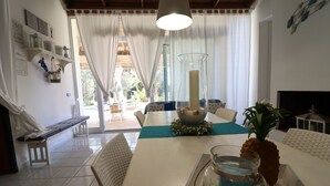 Uma sala de jantar com mesa branca, cadeiras, um vaso e uma tigela de frutas.