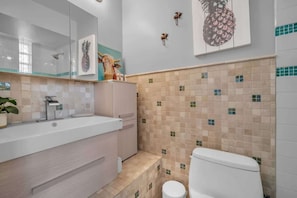 Um banheiro com pia branca, espelho e vaso sanitário. Há azulejos decorativos na parede e uma pequena planta no balcão.