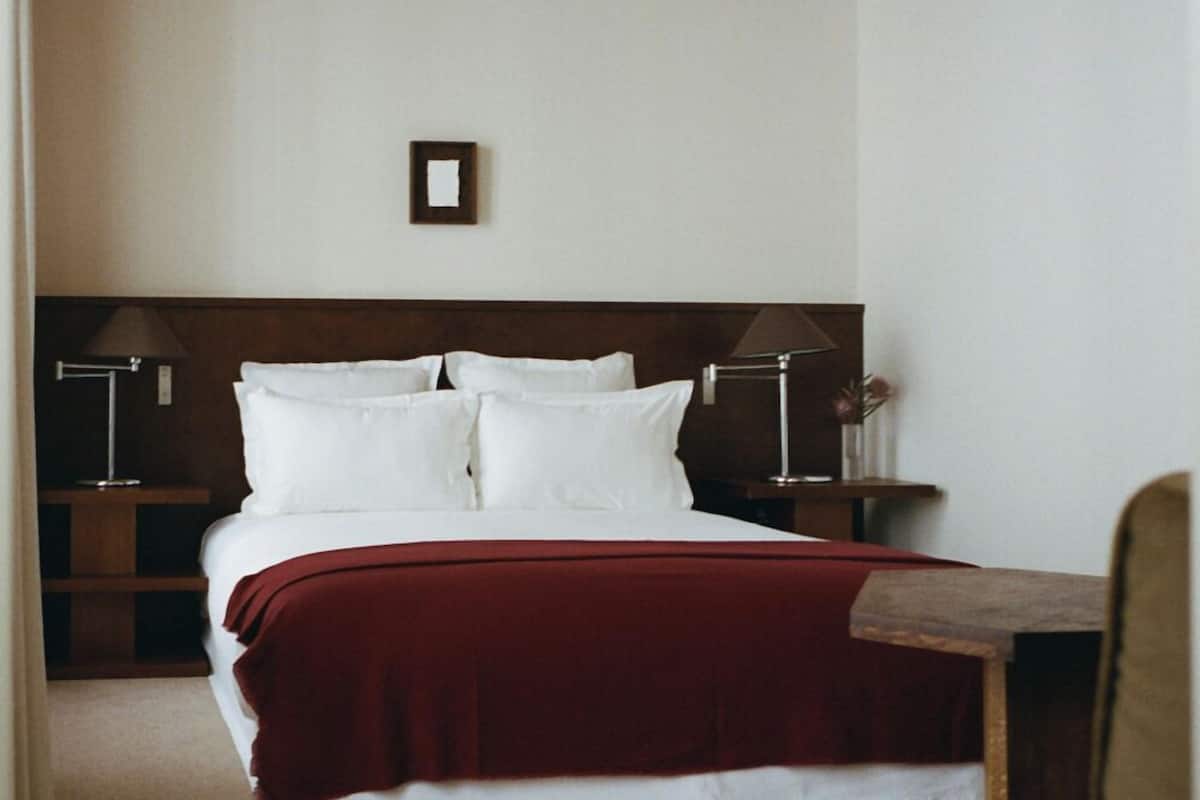 A bedroom with a wooden ceiling, a bed with white bedding and a red blanket, two bedside tables with lamps, and a small picture frame on the wall.