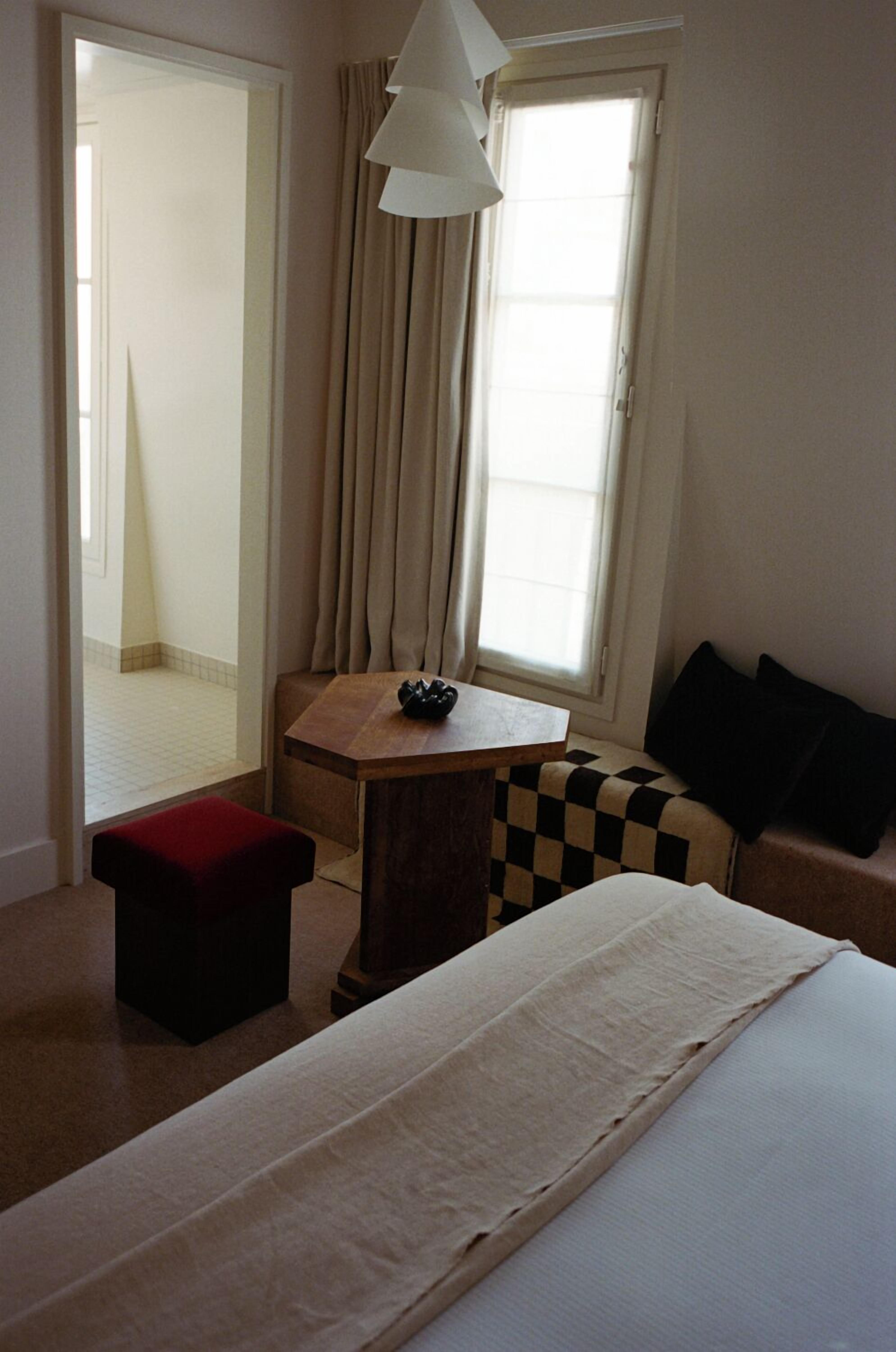 A hotel room with a bed, a small table, a red ottoman, a checkered sofa, a window with curtains, and a hallway leading to another room.