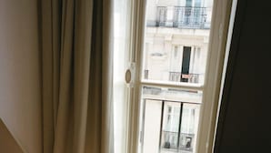 Une pièce avec une seule chaise noire près d’une fenêtre ornée de rideaux, laissant filtrer la lumière du soleil et projeter des ombres sur le sol.