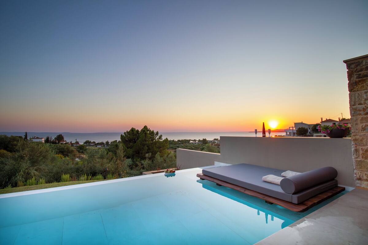 A rooftop infinity pool with a sunset view, a lounge area with cushions, and a stone wall.