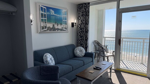 A living room with a sofa, armchair, and a table. There is a painting of a beach scene on the wall.