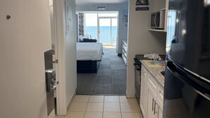 A narrow hallway leading to a bedroom with a view of the sea, a kitchen with a refrigerator and microwave, and a welcome sign.