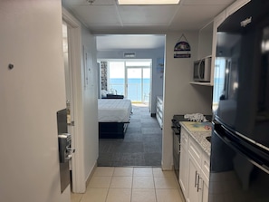 A narrow hallway leading to a bedroom with a view of the sea, a kitchen with a refrigerator and microwave, and a welcome sign.