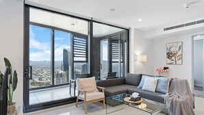 Ein modernes Wohnzimmer mit einer Couch, einem Sessel, einem Glastisch und einer großen Schiebeglastür, die auf einen Balkon mit Stadtblick führt.