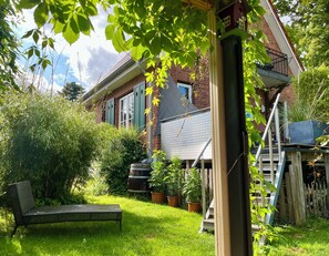 A backyard with a grassy lawn, a metal bench, potted plants, and a brick house with a balcony. - Small country house pearl by the castle park and lake, in dreamlike nature! (Eutin)
