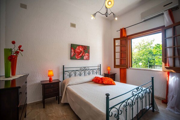 Een slaapkamer met een bed, nachtkastje, lamp, raam met gordijnen en een schilderij aan de muur.