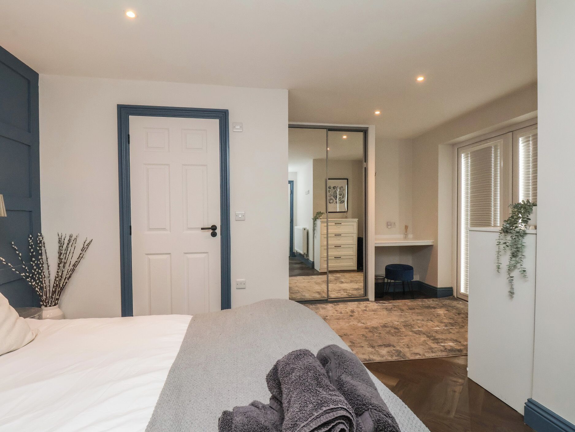 A bedroom with a bed, a door, a mirror, a desk, and a plant.