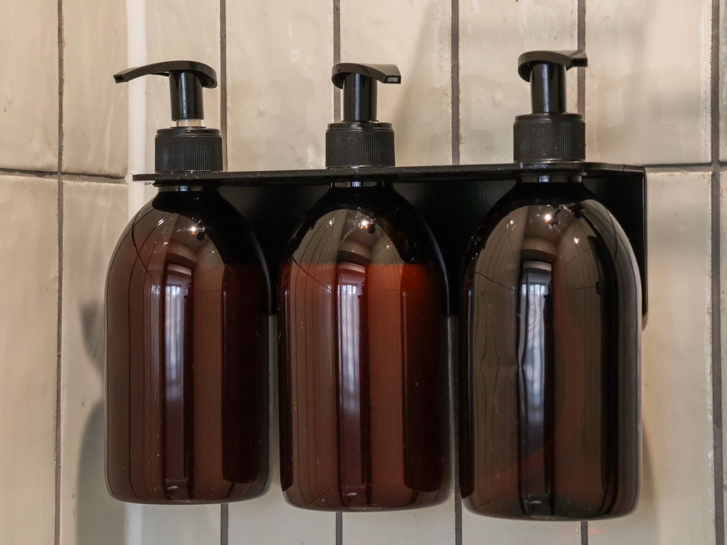 Three brown bottles with black pumps mounted on a black shelf against a tiled wall.