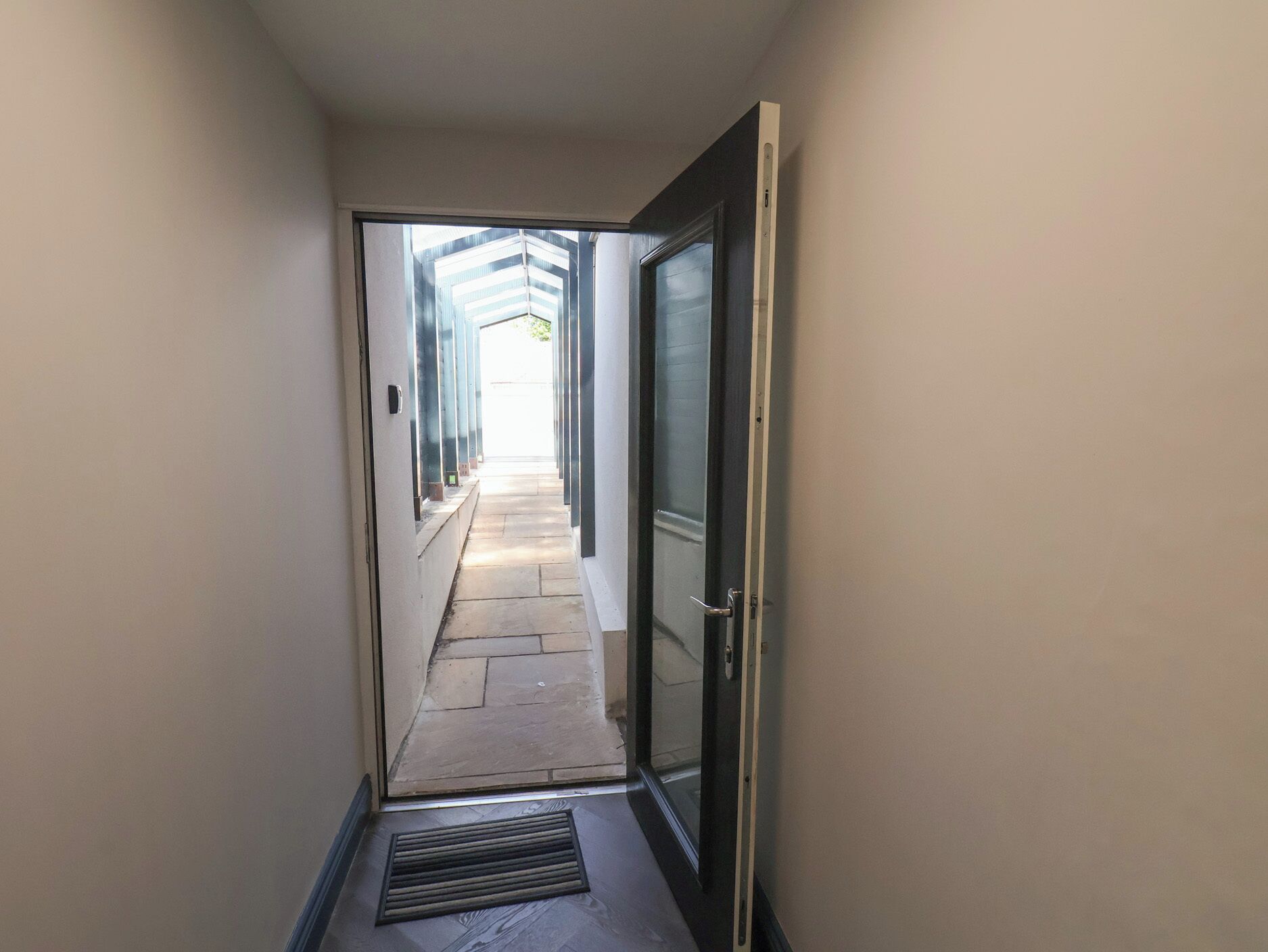 Un couloir étroit mène à une porte ouverte, d’où l’on aperçoit un bâtiment à l’extérieur.