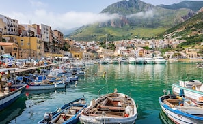 A coastal town with a marina filled with boats, buildings along the waterfront, and a mountainous backdrop.