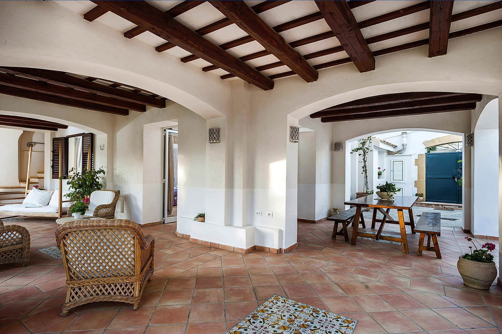 A spacious interior with a high ceiling, exposed wooden beams, and a tiled floor. There are wicker furniture pieces and a table with chairs.