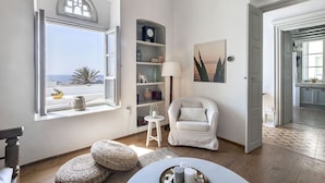 A room with a view of the sea, a white chair, a bookshelf, and a painting.
