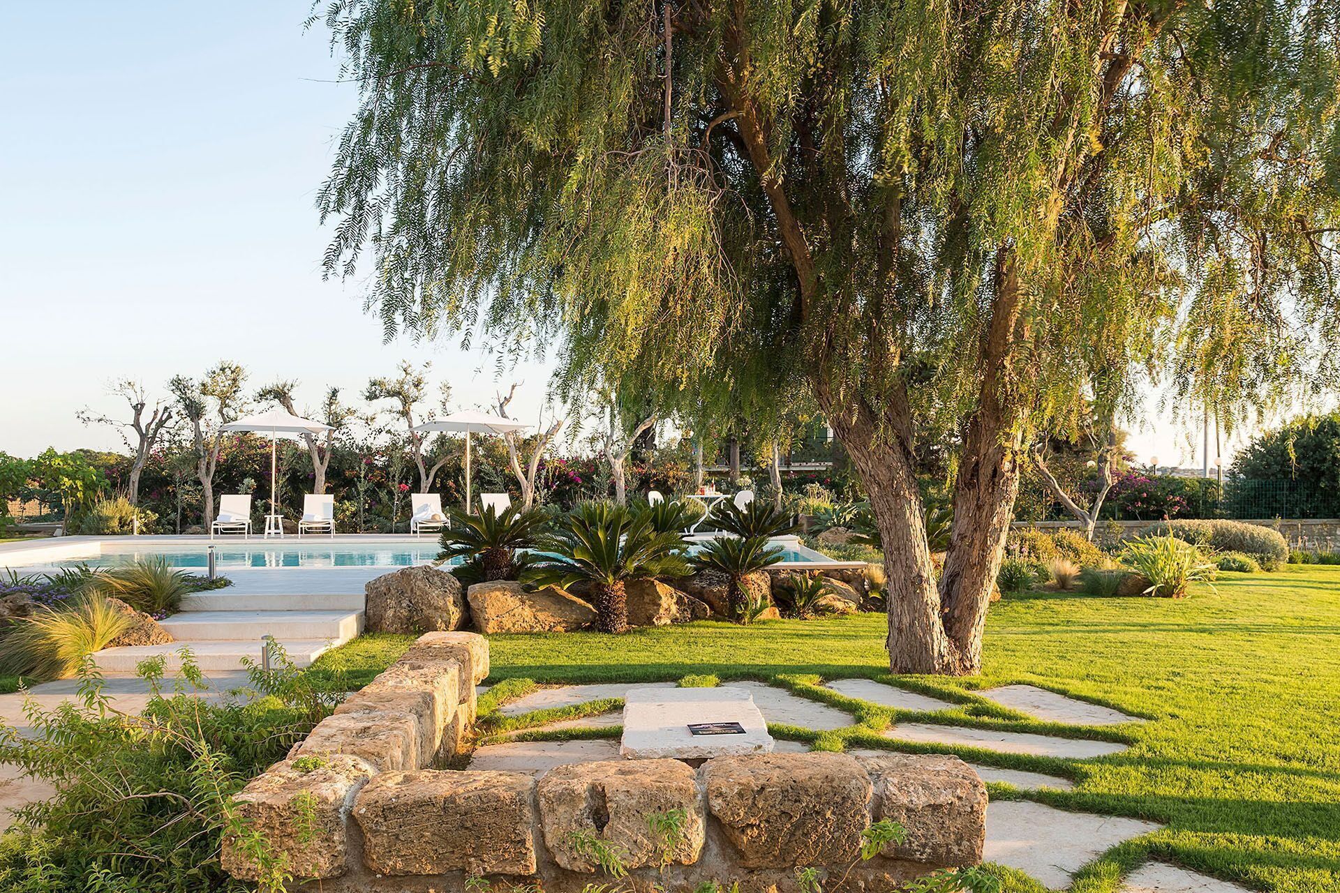 Un área de piscina con un sendero de piedra, sillas de descanso y un árbol grande.