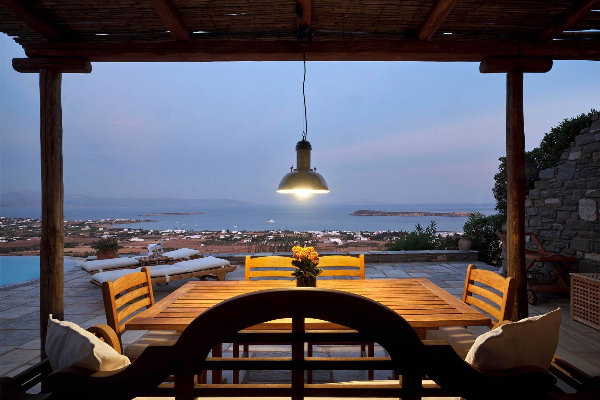 An outdoor dining area with a wooden table and chairs, a hanging light fixture, and a view of the sea and distant land.