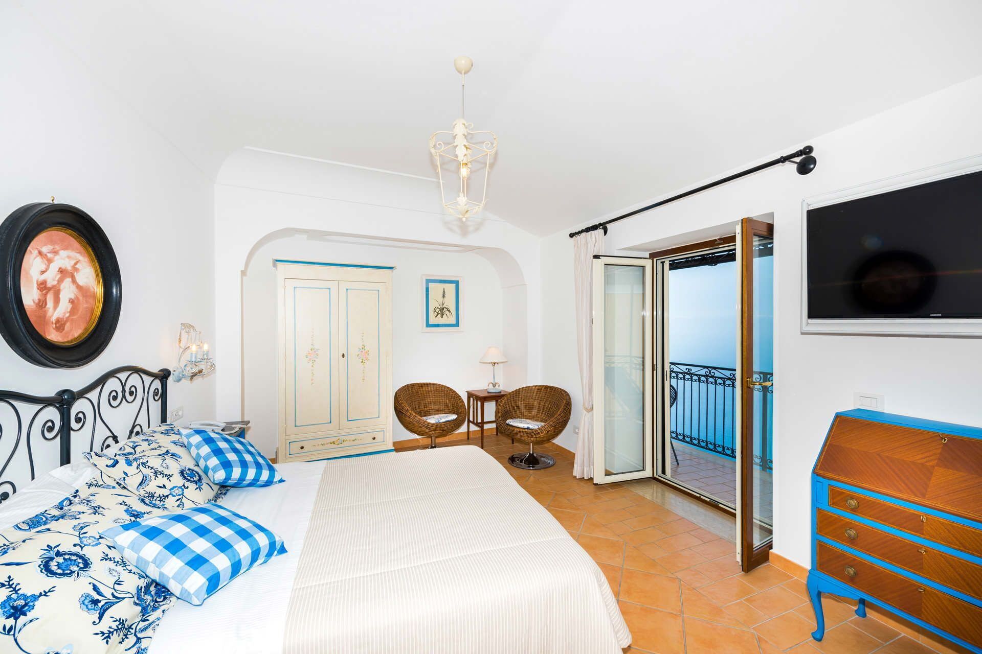 Ein Schlafzimmer mit einem Bett, zwei Korbstühlen, einem kleinen Tisch, einem Kleiderschrank und einer Balkontür.