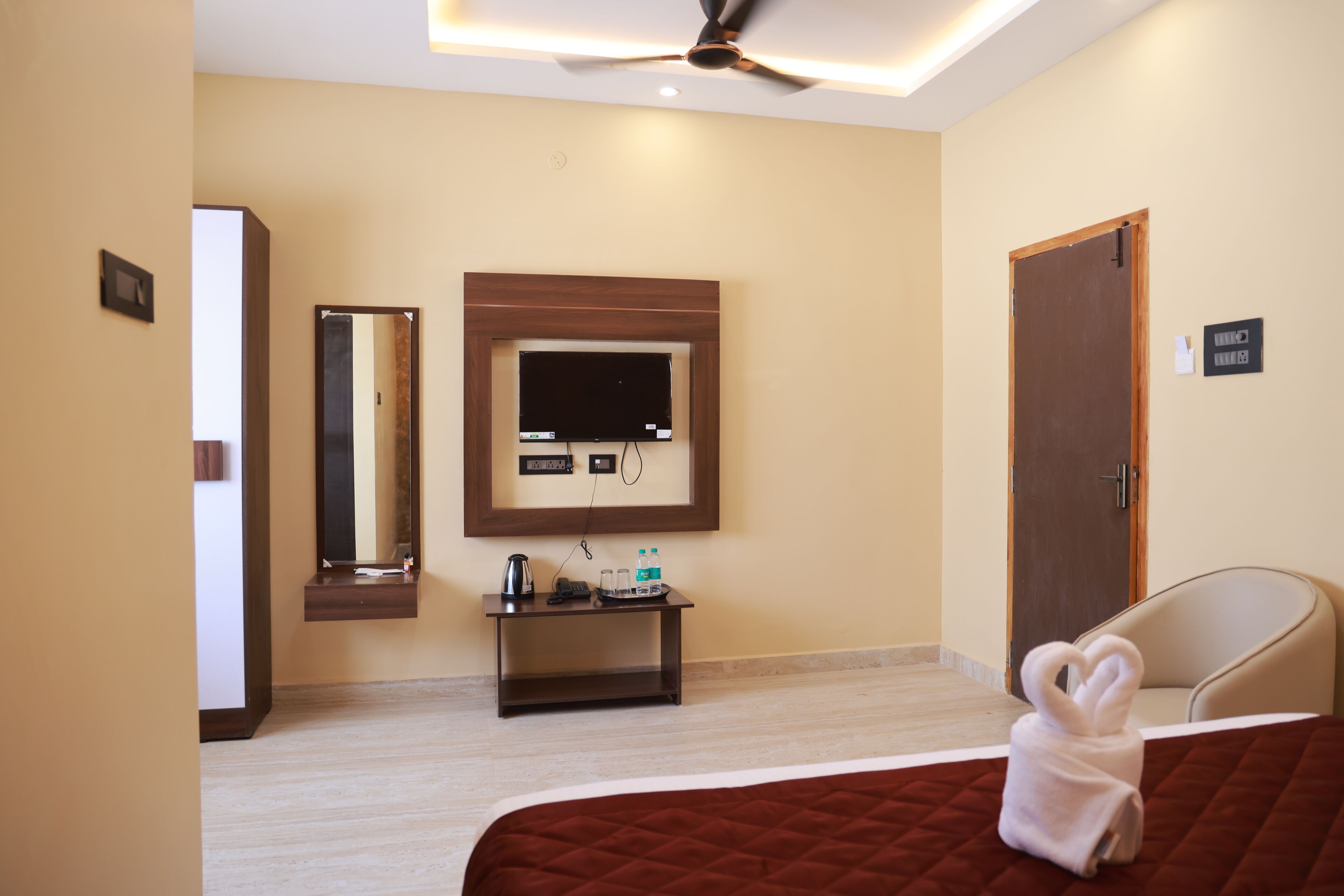 A hotel room with a bed, a TV mounted on the wall, a small table with a kettle and water bottles, a chair, and a door.