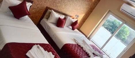 A hotel room with two beds, each with a red quilt and white linens. There is a window with a view of trees and a ceiling fan.