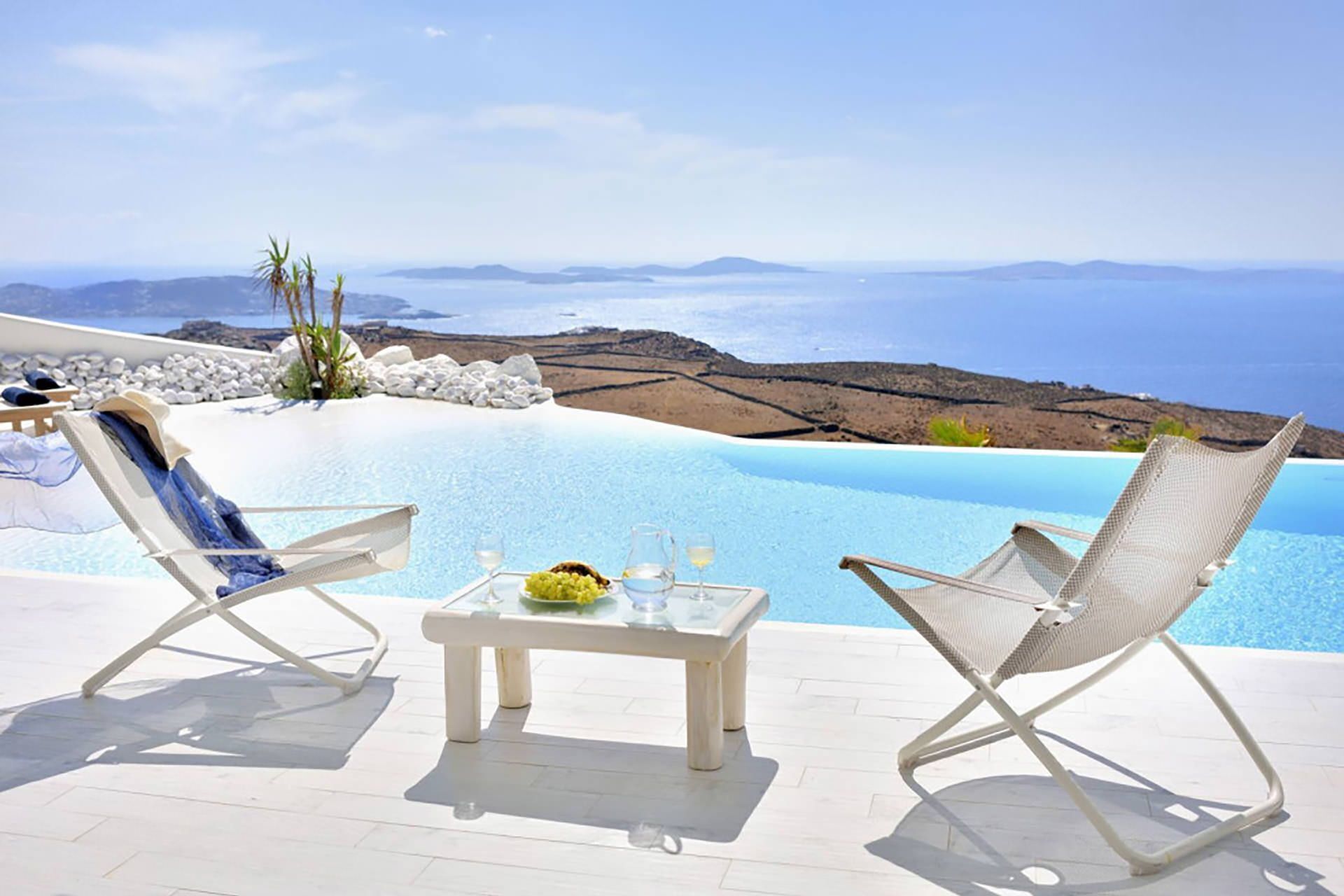 A poolside area with two lounge chairs, a table with a glass of wine, and a view of the sea and landscape.