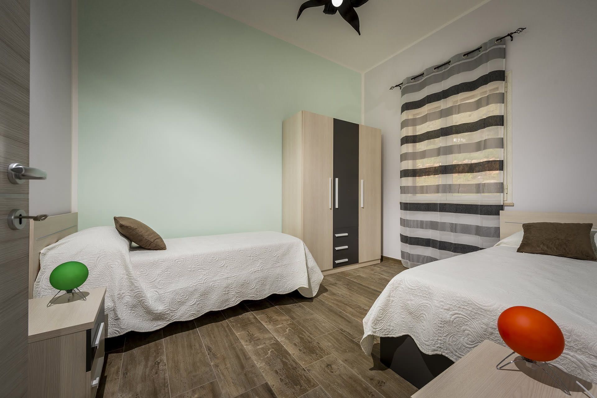 Quarto com duas camas, um guarda-roupa e uma janela com cortina listrada.