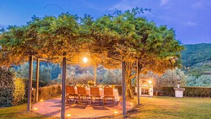 Ein Essbereich im Freien mit einer Pergola, Tischen und Stühlen, umgeben von Grün und Blick auf Hügel.