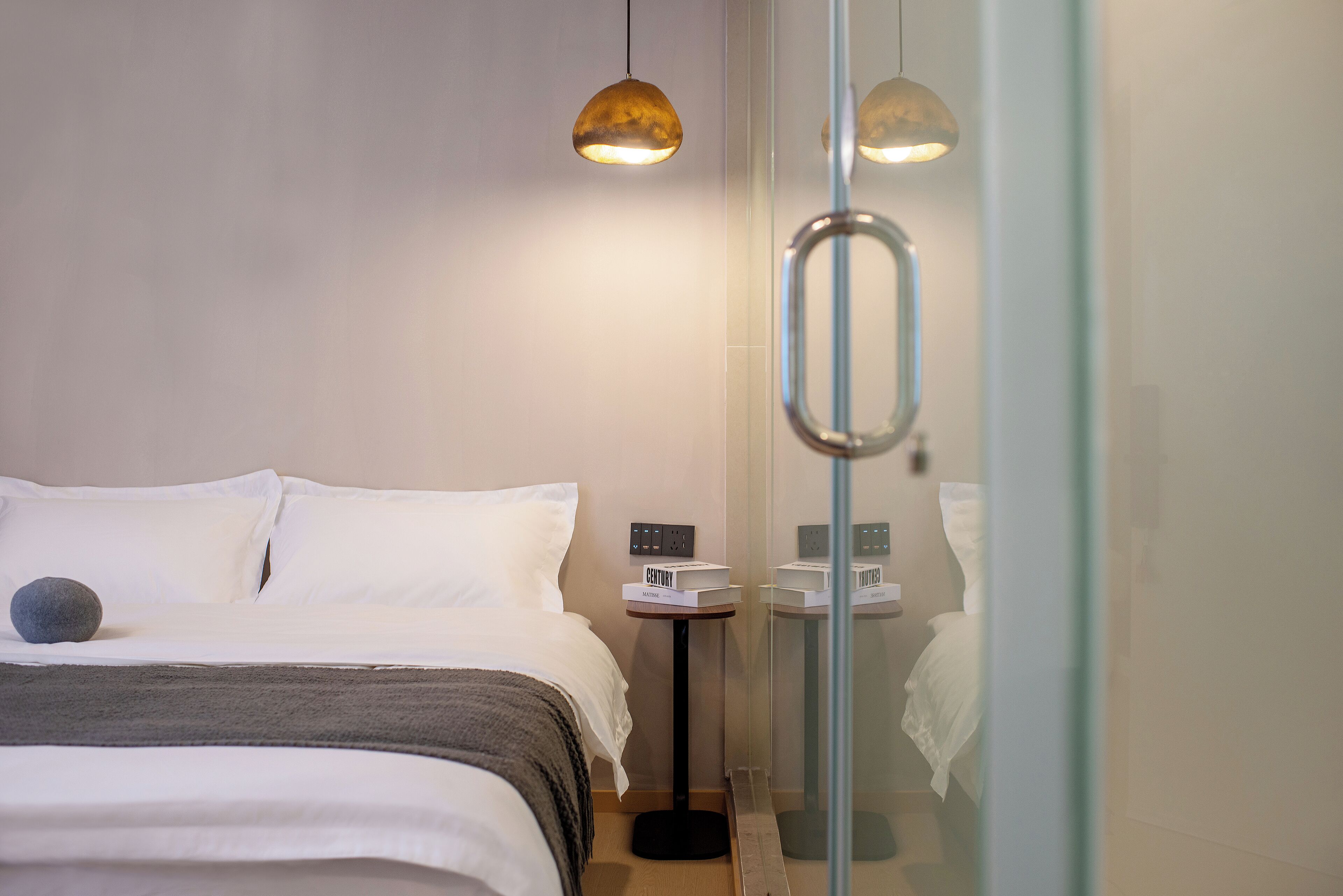 A modern hotel room with a neatly made bed, a small bedside table with a lamp, and a glass shower enclosure.