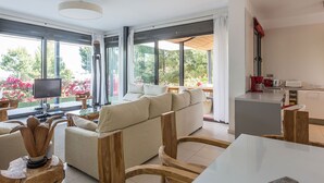 Un salon moderne avec un canapé, une table basse et un téléviseur. Il y a un coin cuisine équipé d’un réfrigérateur et d’un appareil électroménager rouge. De grandes fenêtres offrent une vue sur la verdure à l’extérieur.