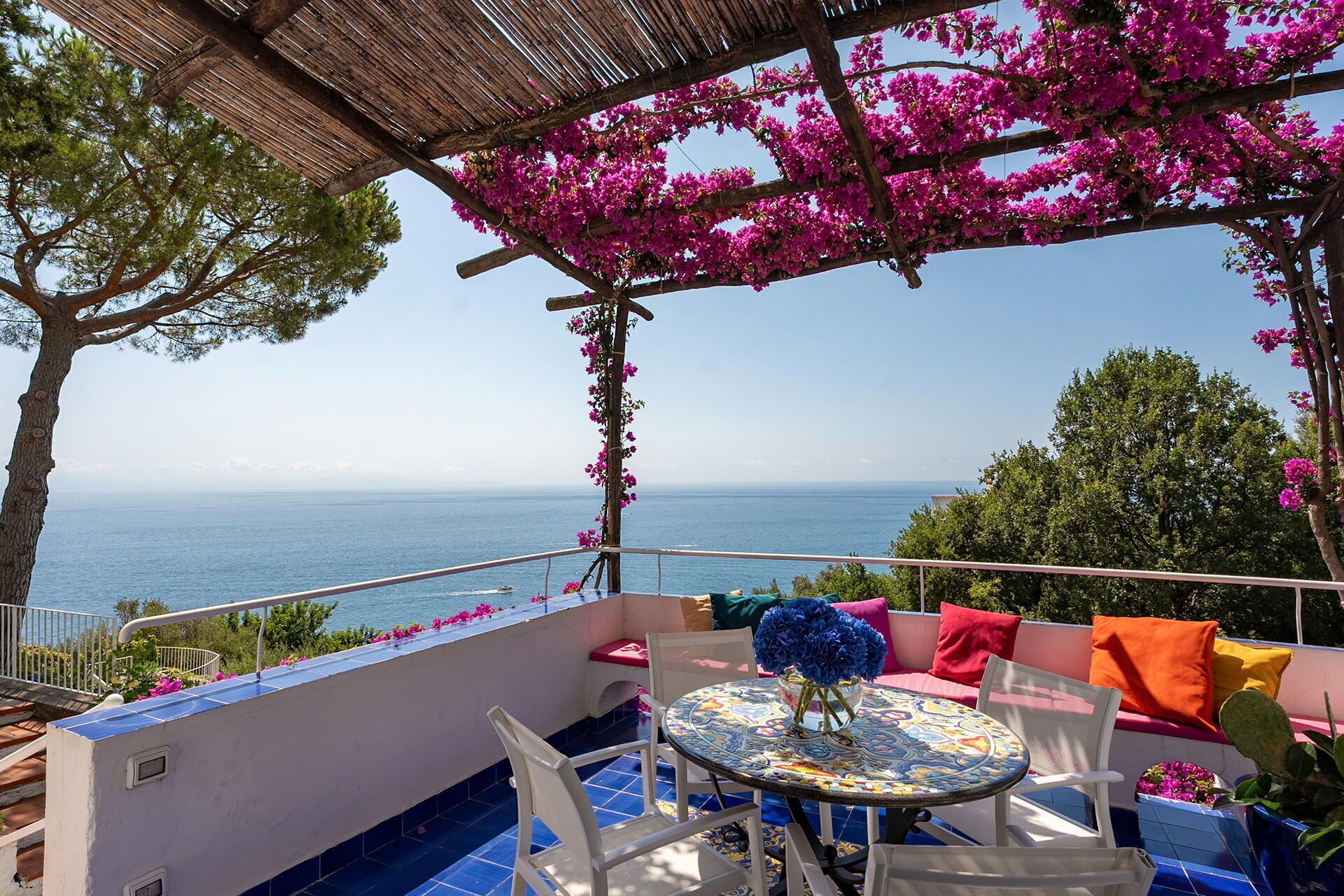 A terrace with a view of the sea, a table with a vase of flowers, and colorful cushions.