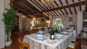 A dining room with a table set for six, featuring a lace tablecloth, candles, and a bottle of wine. The room has wooden beams on the ceiling and a large potted plant.