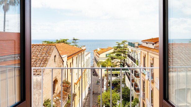 A narrow street with buildings on either side, a clear view of the sea, and a balcony with a metal railing. - Interno Sorrentino with sea view - My Rental Homes (Sorrento)