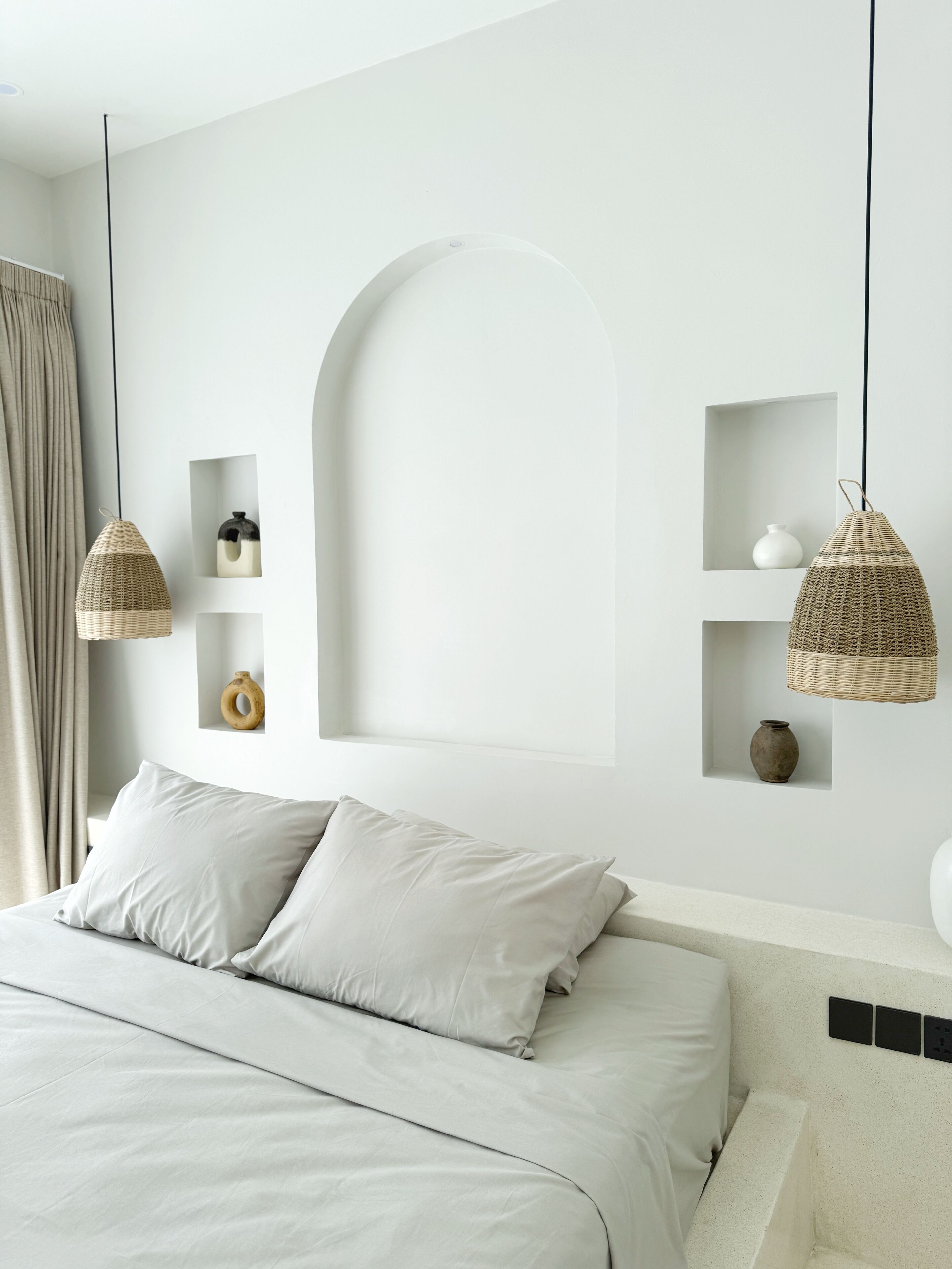 Un dormitorio minimalista con cama, dos lámparas colgantes de mimbre y una pared blanca con estantes empotrados que exhiben objetos decorativos.