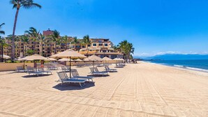Une plage avec des transats et des parasols, un bâtiment d’hôtel en arrière-plan et un ciel bleu dégagé.