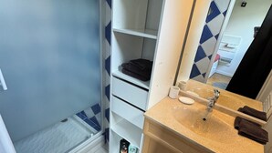 Une salle de bain avec une douche, un lavabo et un miroir. On y trouve des étagères avec des articles de toilette et un tapis noir sur le sol.