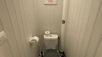 Un baño pequeño y estrecho con paredes blancas y un inodoro. Hay un rollo de papel higiénico en la pared y un estante pequeño con una botella y un cepillo.
