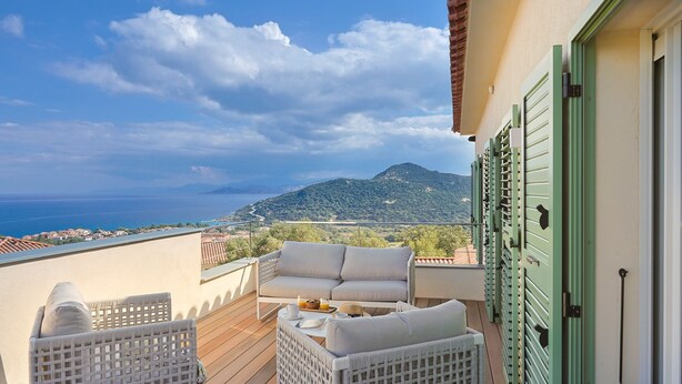 A balcony with a sofa and chairs, a table with a tray of items, and a view of the sea and mountains.