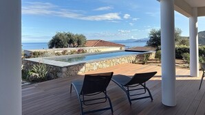 A poolside area with two lounge chairs, a stone wall, and a view of the sea and mountains.