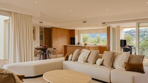 A modern living room with a large sectional sofa, a dining area, and a kitchen with a view of the outdoors.