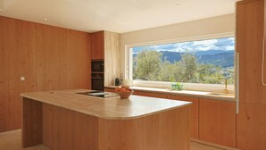 A modern kitchen with a large island, wooden cabinetry, and a large window offering a view of mountains and trees.