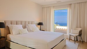 A large bed with white linens, a nightstand with a lamp, a chair, and a view of the sea through a balcony.