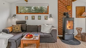 Un salon chaleureux avec un canapé gris, une table basse en bois sur laquelle sont disposés des fruits, un mur en briques et un poêle à bois.
