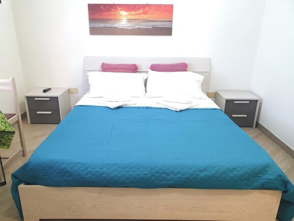A neatly made bed with a blue quilt, two white pillows, and a pink throw. There are two bedside tables with drawers, one on each side of the bed. A painting of a sunset over the ocean hangs above the bed. - Studio Apartment - La Ciaccia Style (La Ciaccia)